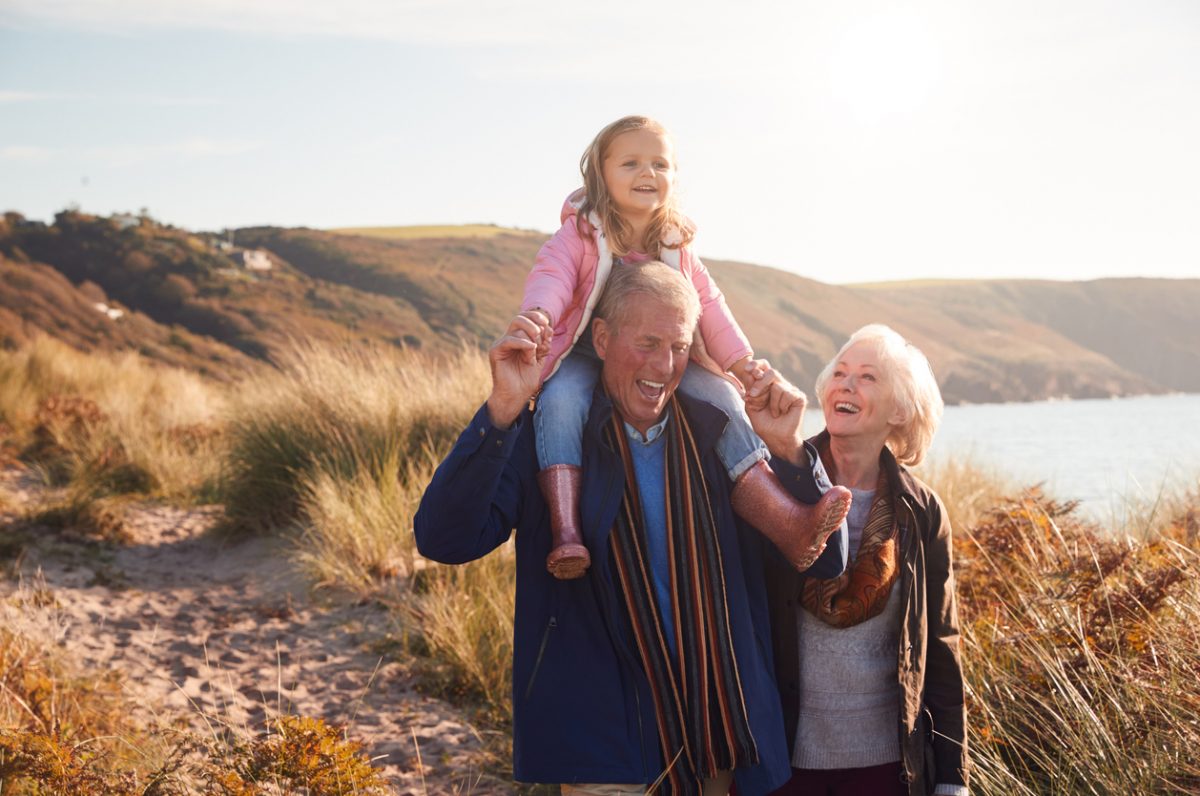 Grandfather carrying granddaughter on shoulders walking along coastline with grandma beside them