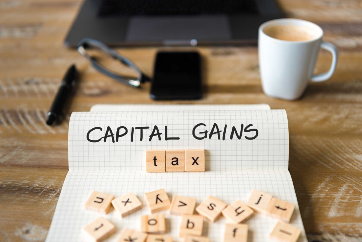 Graphic of a piece of paper with the words 'Capital Gains' handwritten on the page and wooden squares with letters on scattered across. The word 'tax' is spelled out in the wooden squares beneath 'Capital Gains'.